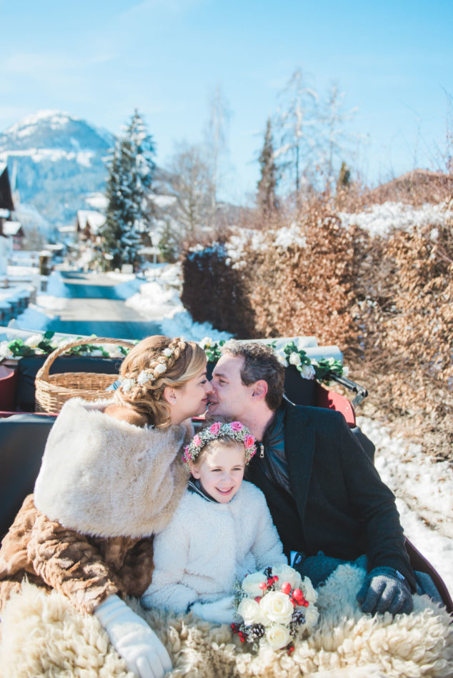 A bride, groom and their daughter in a sleigh during a photo shoot on their wedding day in Austria
